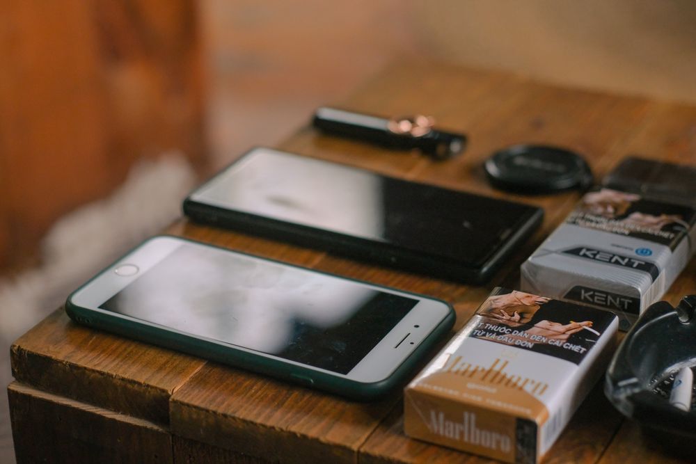 Close-up of mobile phones, cigarette packs, and gadgets on a wooden table.