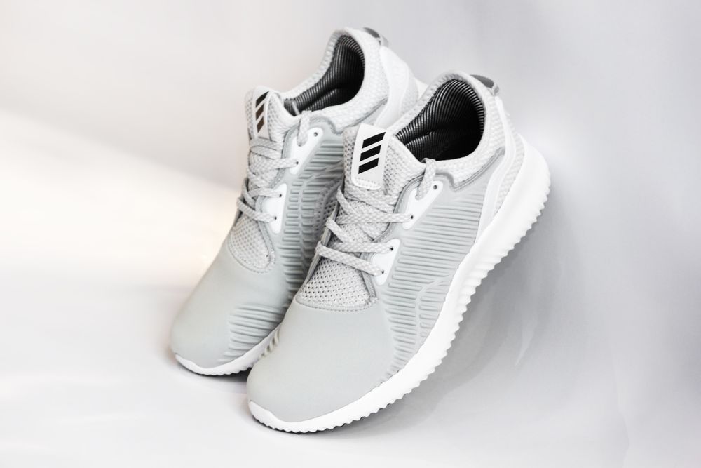 Pair of sports shoes with gray dense texture of surface and rubber inserts from sided and laces and white soles placed with heels up leaning on glance gray background