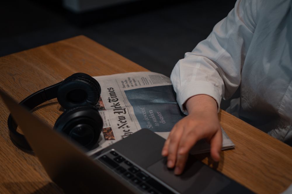 A person works on a laptop with headphones and a newspaper on a wooden table.
