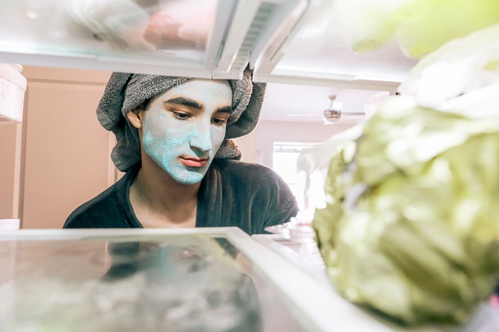 A man with a blue face mask looks into the refrigerator, towel wrapped on head, in a bright kitchen.