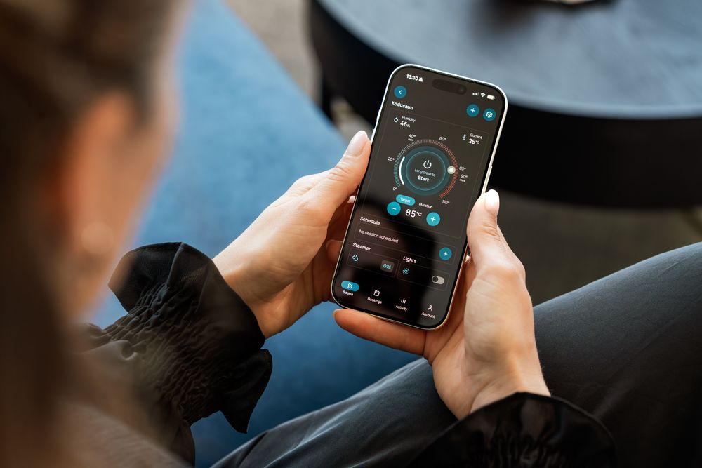 A woman controls a smart sauna system via a touchscreen app on her smartphone indoors.