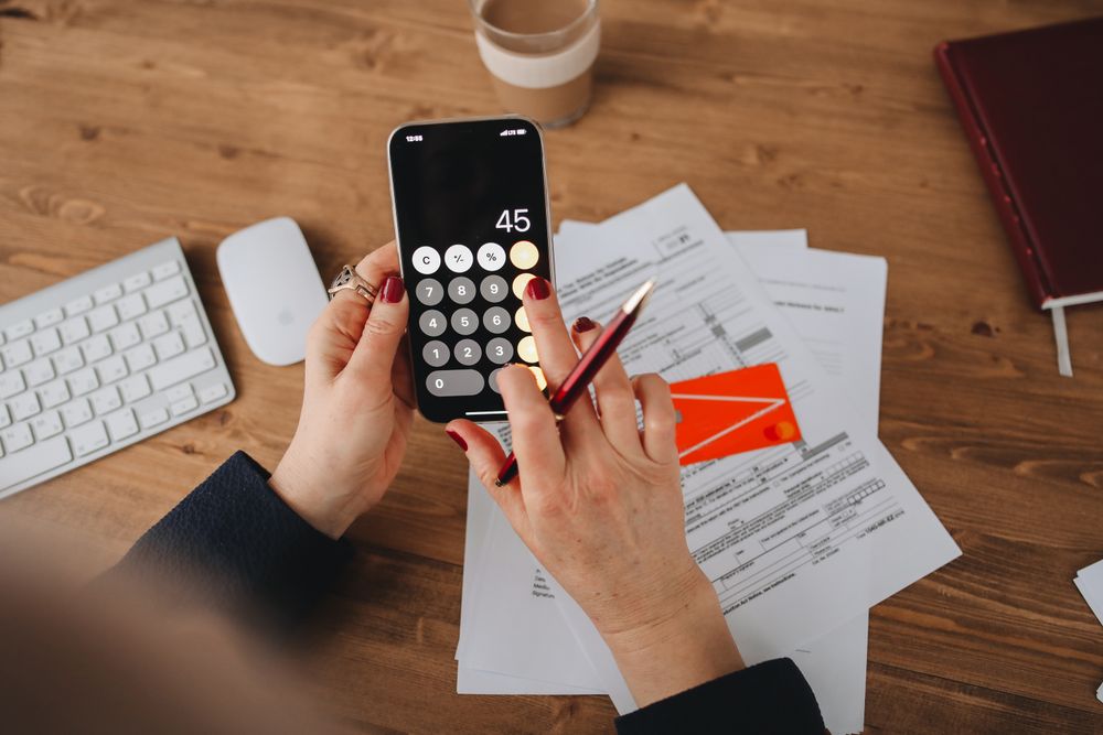 Hands using a calculator app on a smartphone over financial documents. Perfect for illustrating finance or technology themes.